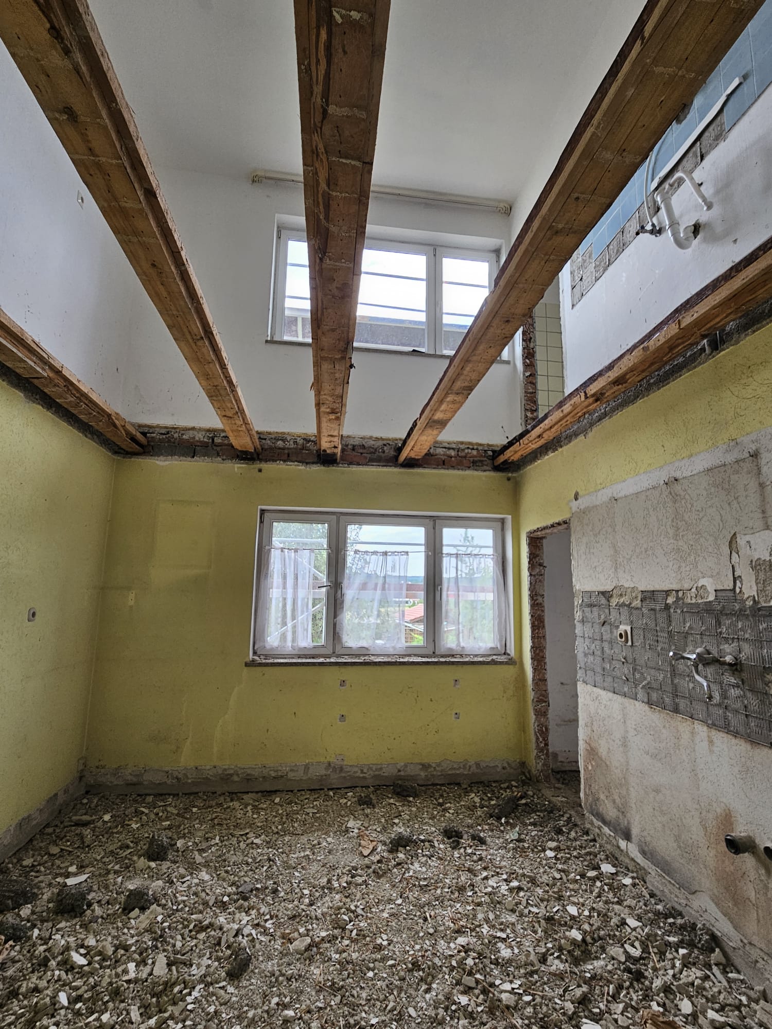Raum im Rohbau: offene Holzbalkendecke, abgeschlagene Fliesen, Schutt am Boden